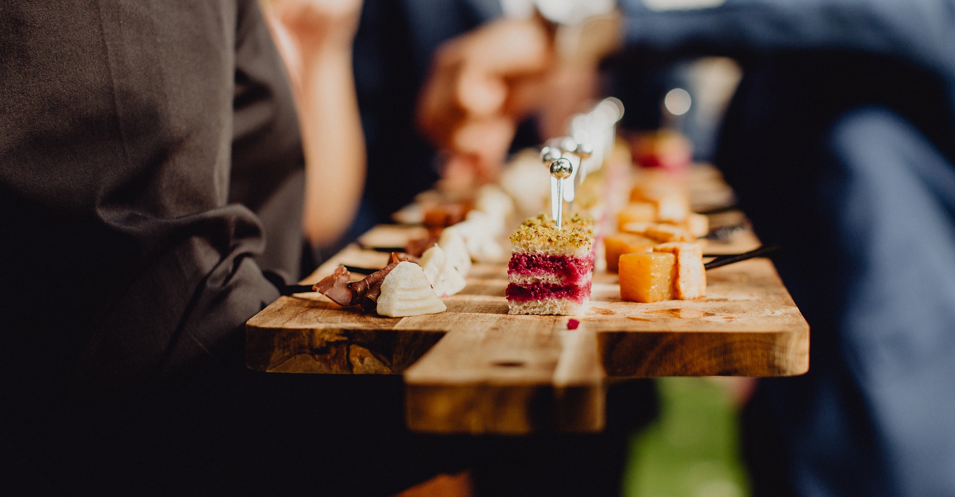 Personne en costume noir servant un plateau en bois d'amuse-gueules variés : charcuterie, fromages et canapés élégants lors d'un événement.