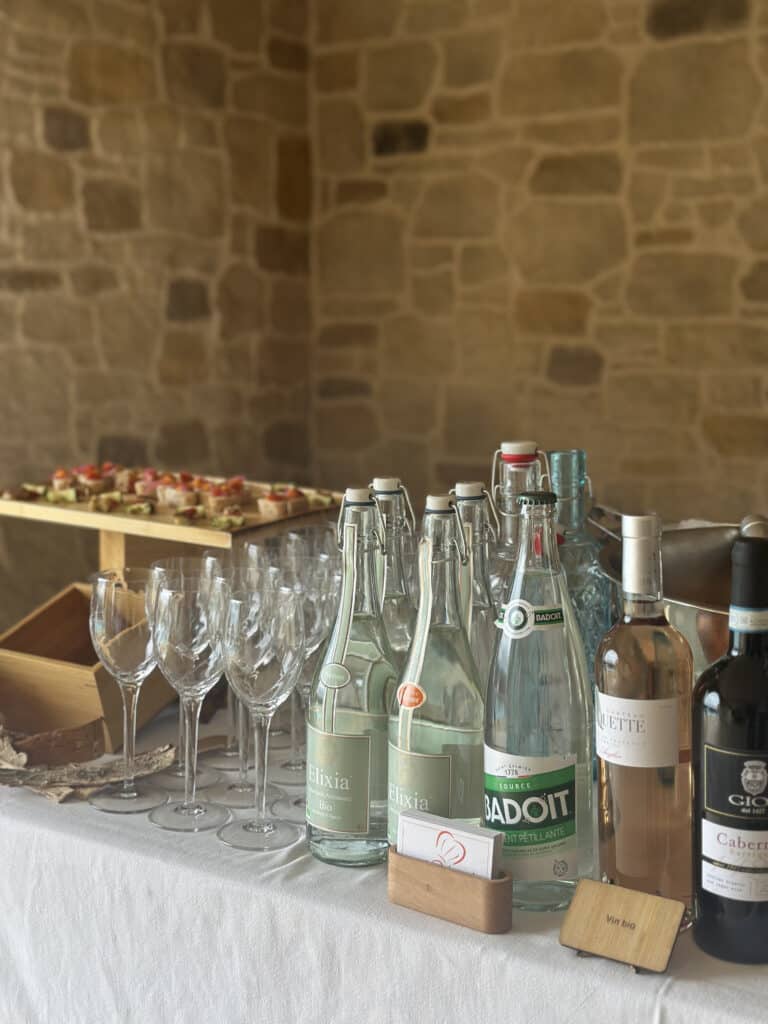 Buffet apéritif avec bouteilles d'eau Elixia & Badoit, vins Château Quette & GIO Cabernet bio. Canapés, verres, et mur en pierre.