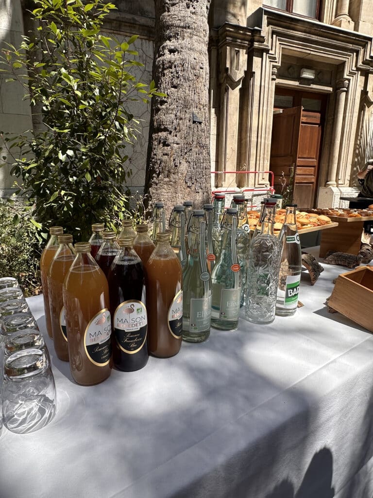 Table de buffet extérieur avec jus Maison Benedetti (Pomme Bio, Framboise), eaux Elixia, Badoit et verres. Façade historique et arbres en arrière-plan.