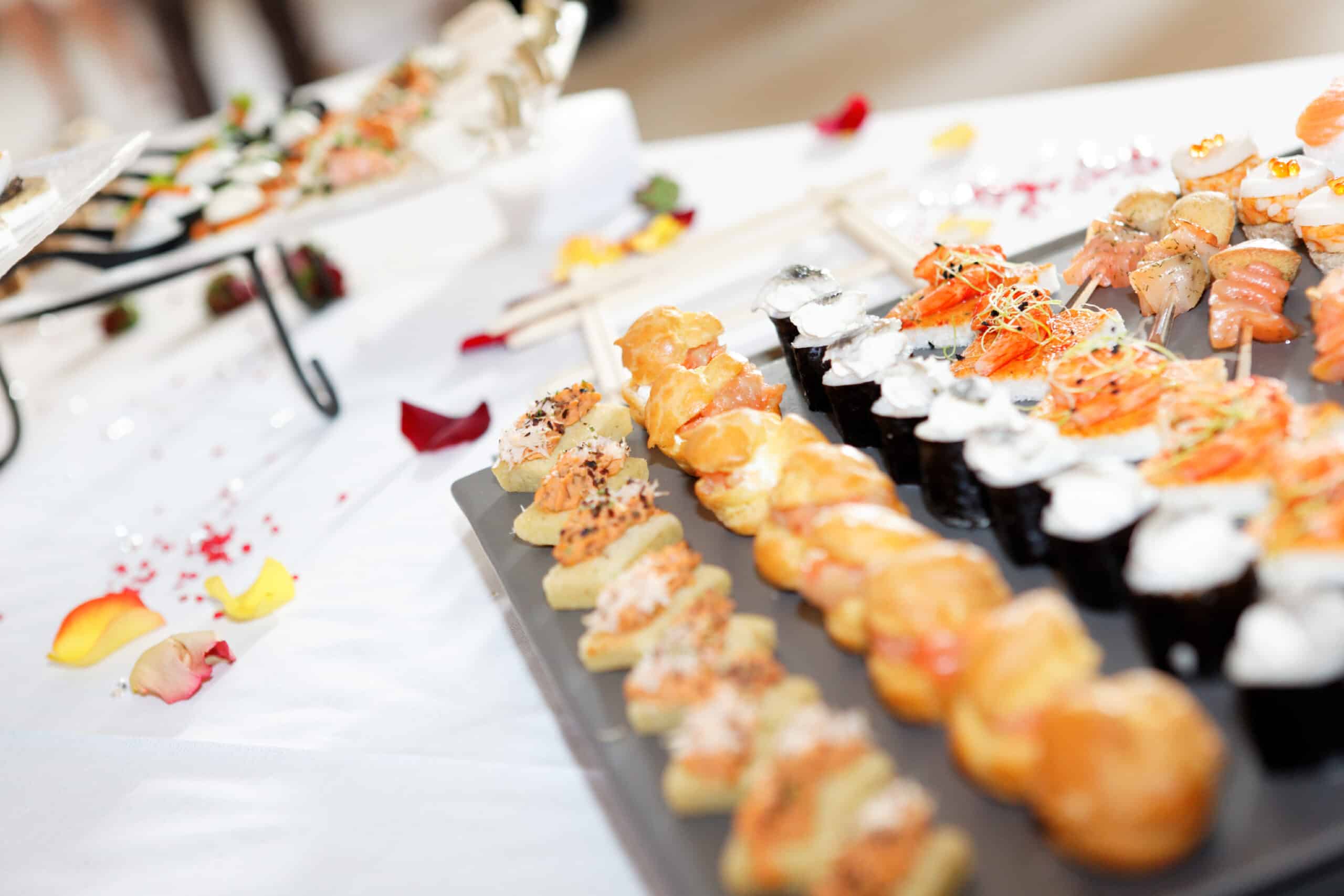 Buffet de réception élégant avec canapés variés, sushis frais et amuse-bouches gourmands sur table blanche décorée de pétales.