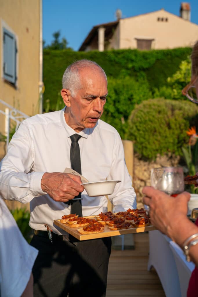 Serveur élégant en chemise blanche et cravate noire proposant des toasts apéritifs sur un plateau en bois lors d'une réception extérieure.