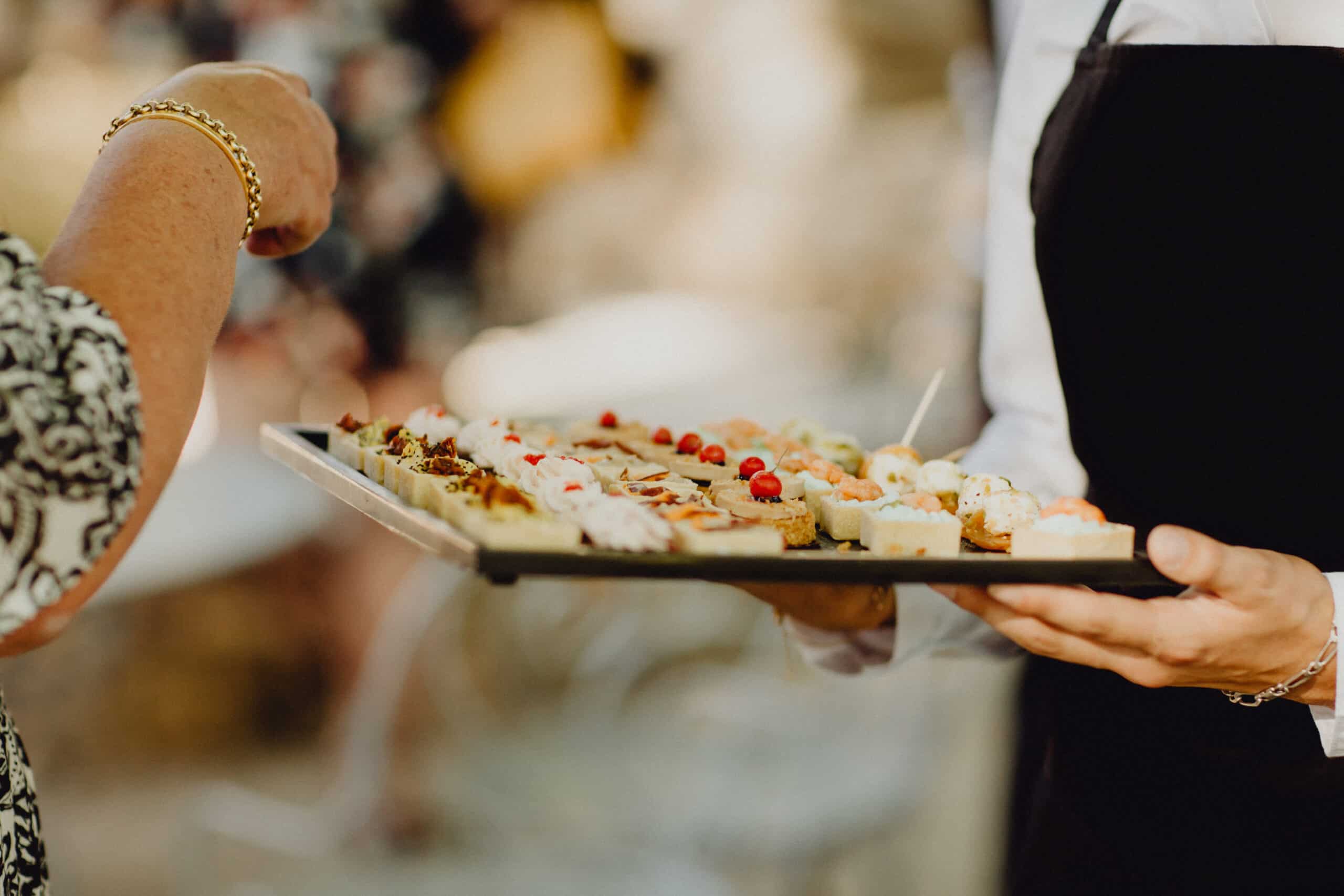 Serveur présentant un plateau d'amuse-gueules variés à une invitée portant un bracelet doré. Service traiteur événementiel.