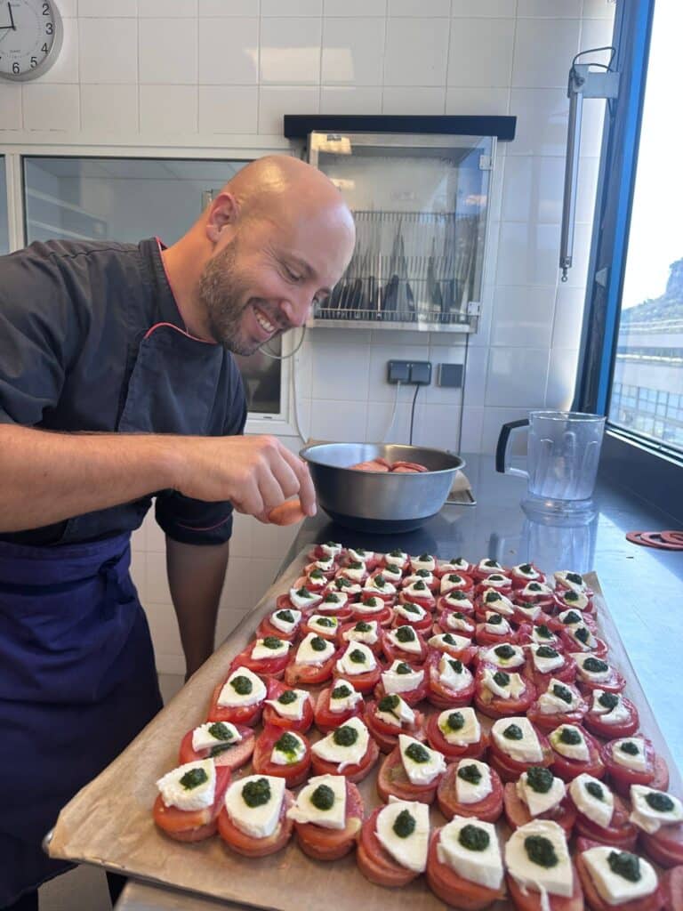 Chef souriant préparant des amuse-bouches type bruschetta tomate mozzarella basilic sur un plateau, horloge murale indique 5 heures.